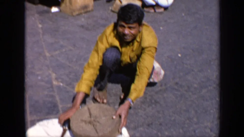 In the 1970 India home-movie frame, a crouching man holds a round stone object while working or performing on the pavement.
