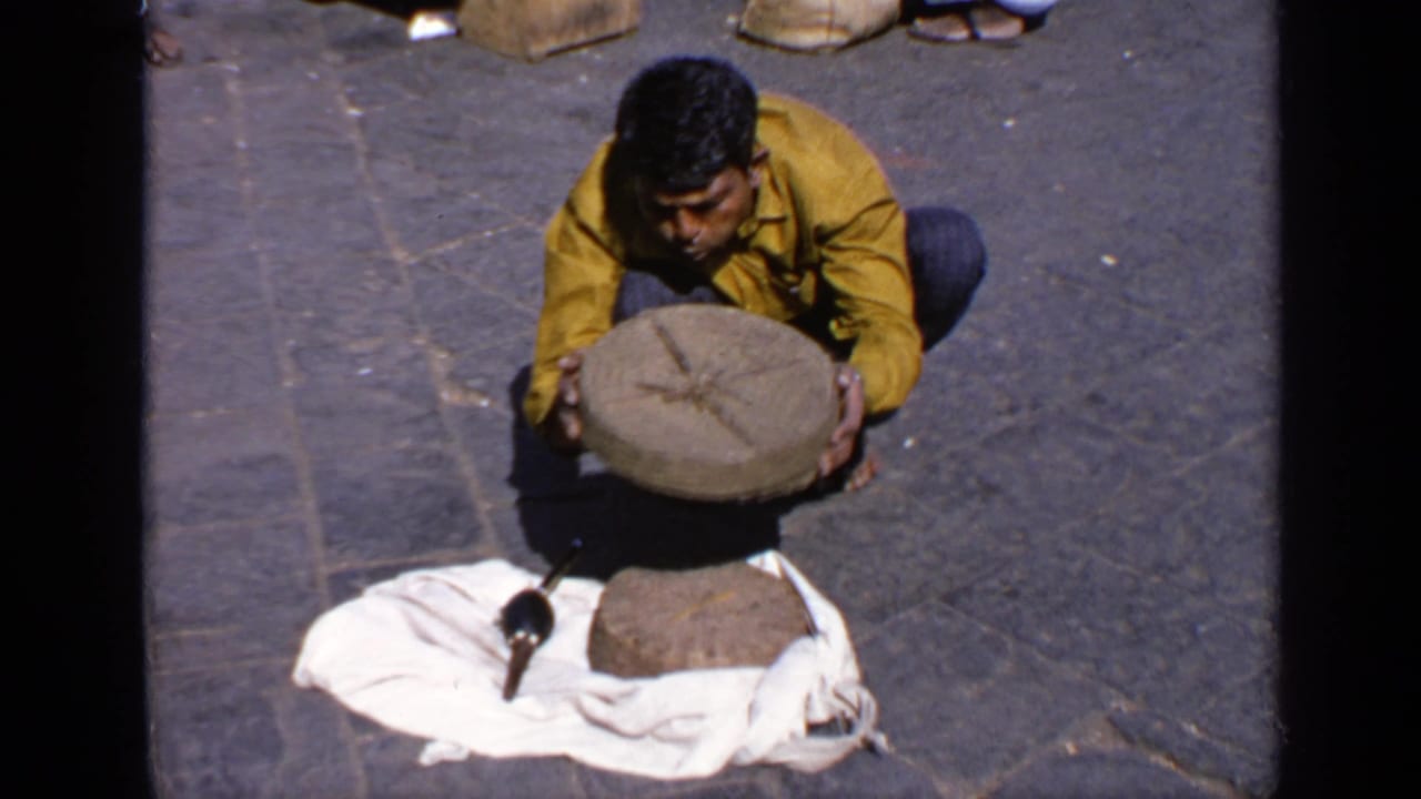 Crouching man lifts a round lid beside a cloth-covered basket on stone pavement.