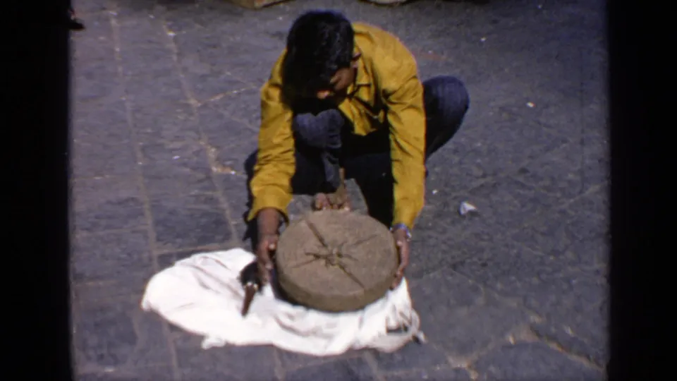 The crouching performer positions the round lid over the basket area on a stone-paved outdoor surface in India.