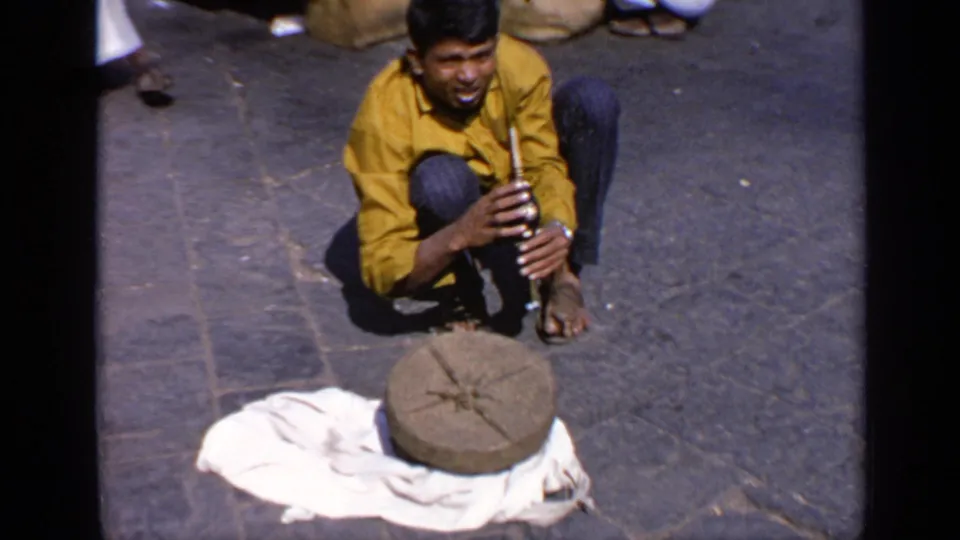 The performer crouches with a wind instrument in hand, with the round lid and cloth-covered basket on the pavement before him.