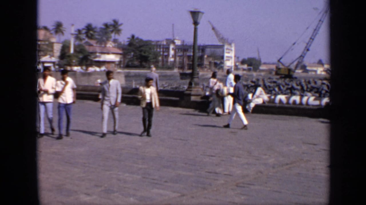 Men walking and standing on a waterfront pier with cranes and buildings beyond.