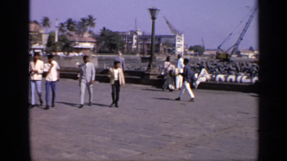 1970 India home-movie frame showing men on a stone pier beside a harbor with cranes, buildings, and palm trees in the background.
