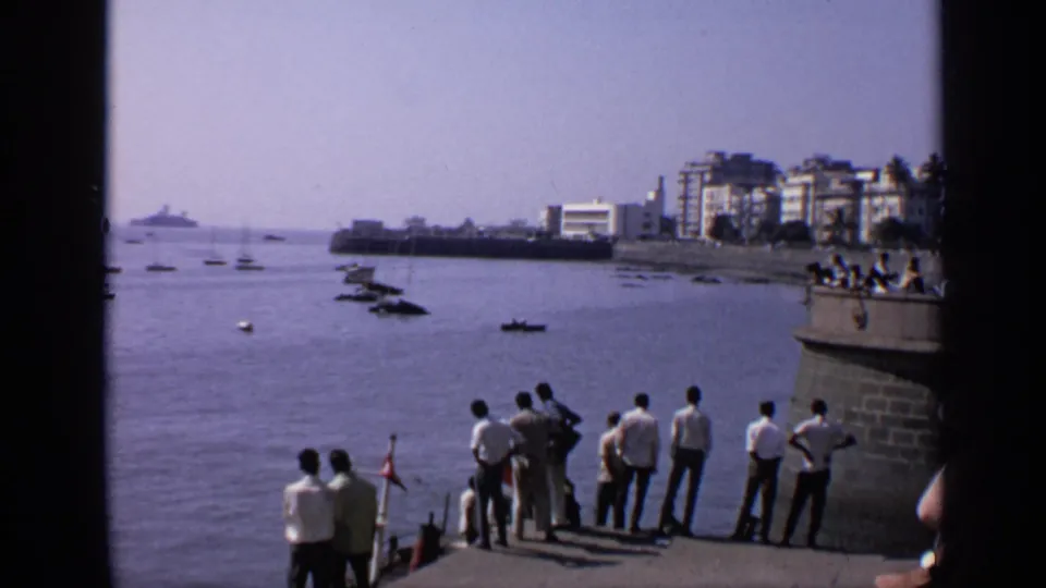 1970 India waterfront view with men gathered at a pier edge, small boats on calm water, and buildings along the far shore.