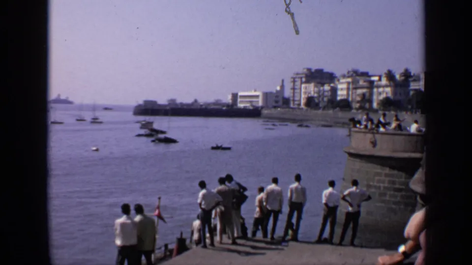 1970 India home-movie frame of men watching a harbor from a stone pier, with boats, buildings, and a distant vessel visible.