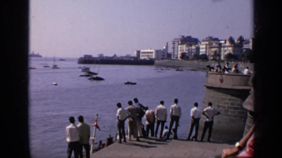 1970 India waterfront frame showing men gathered at a pier overlook, boats on the harbor, and mid-rise buildings along the shore.