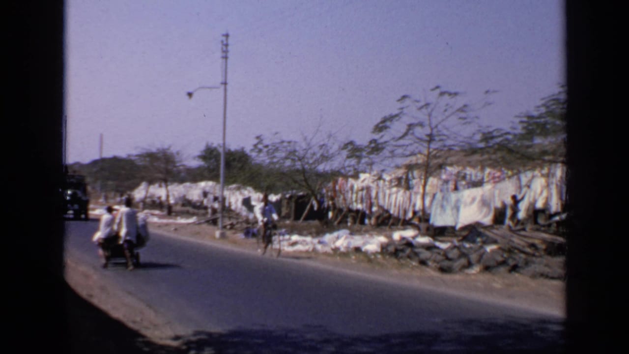 1970 India home-movie frame showing bicyclists, pedestrians, a road edge, hanging white sheets, and sparse roadside trees.