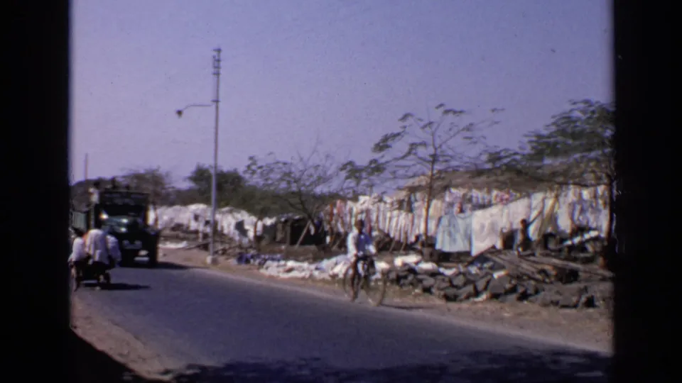 1970 India street frame with a truck, a bicyclist, pedestrians, hanging sheets, and roadside work areas.