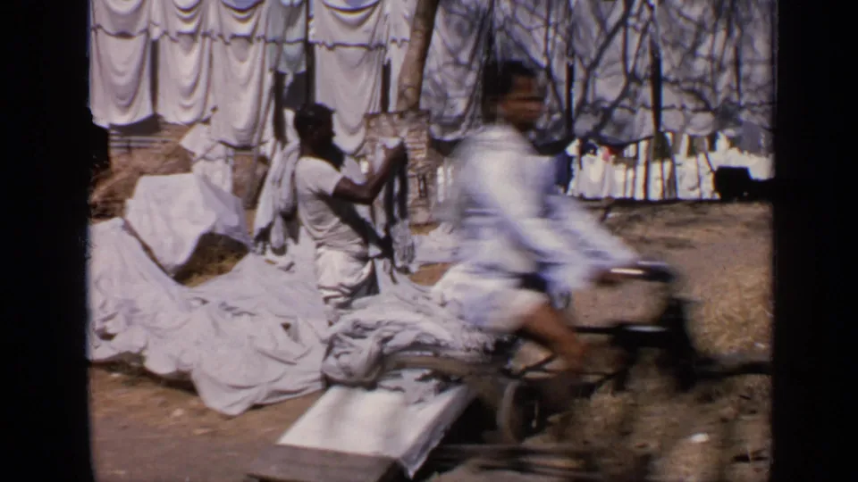1970 India home-movie frame showing a man by laundry, a low cart, hanging sheets, and a blurred passing bicyclist.