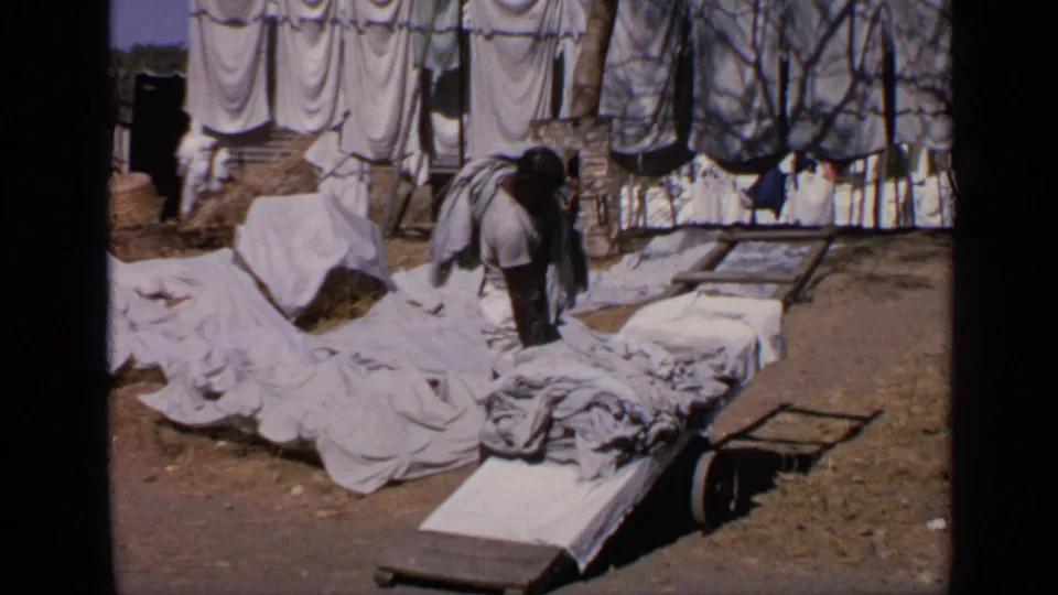 1970 India roadside frame showing a man sorting or handling white sheets near a low cart and laundry lines.