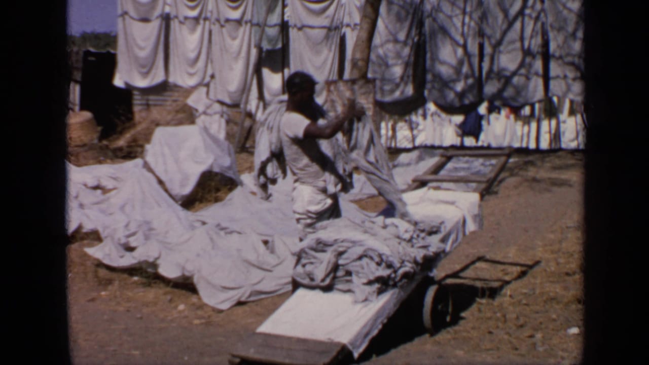 In India around 1970, a man stands near wooden tables covered with white laundry while more garments hang to dry behind him.