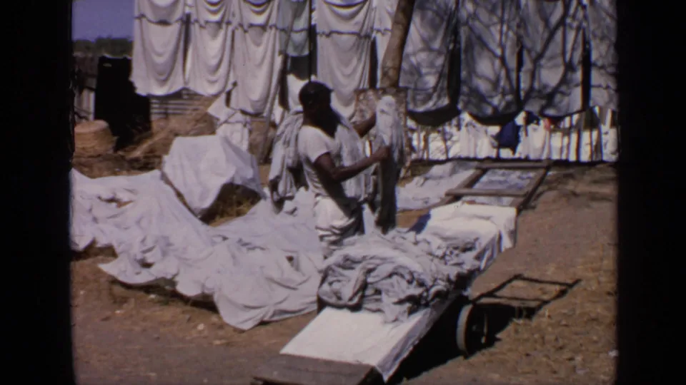 The man carries laundry beside low wooden tables, with white sheets and darker garments hanging on lines in the background.