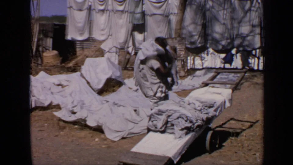 The man bends over or gathers cloth near a broad spread of white laundry, with drying lines and simple yard structures behind him.