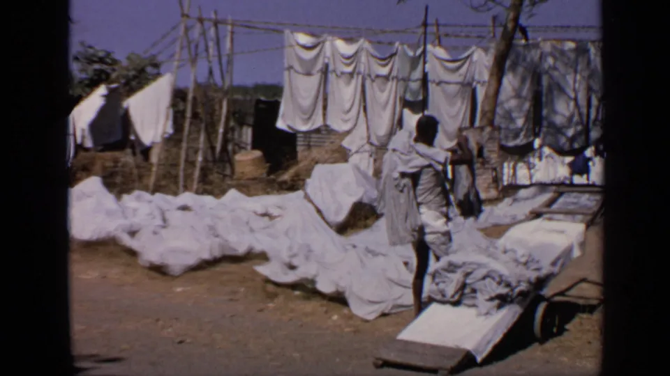 A man carries a large cloth bundle over his shoulder in an outdoor Indian laundry area, with linens and garments drying behind him.