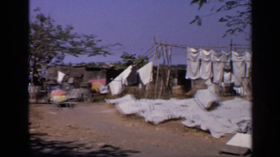 1970 India home-movie frame with motion blur near a dirt compound where sheets hang from wooden supports beside low shelters.