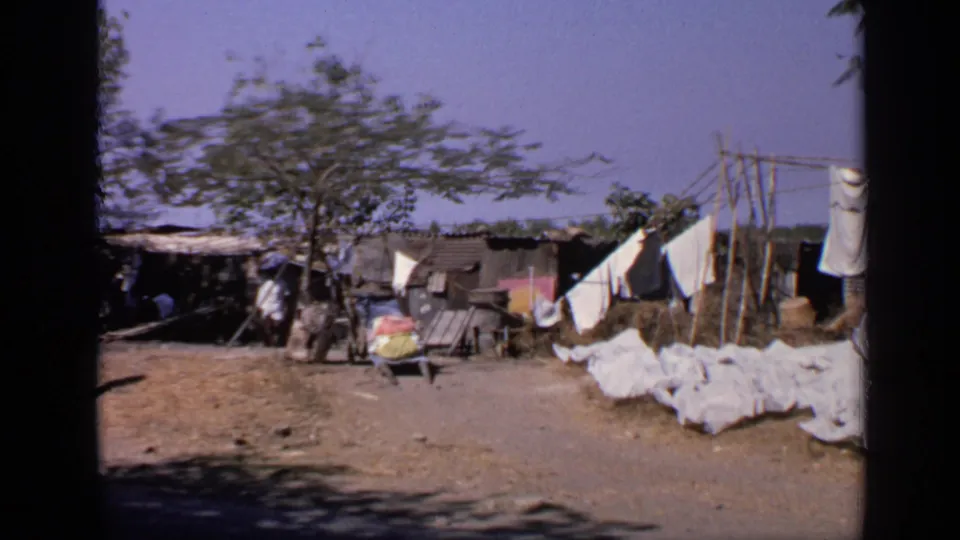 1970 India 8mm frame looking across a dirt camp area with white fabric drying on lines and ground near shaded shelters.
