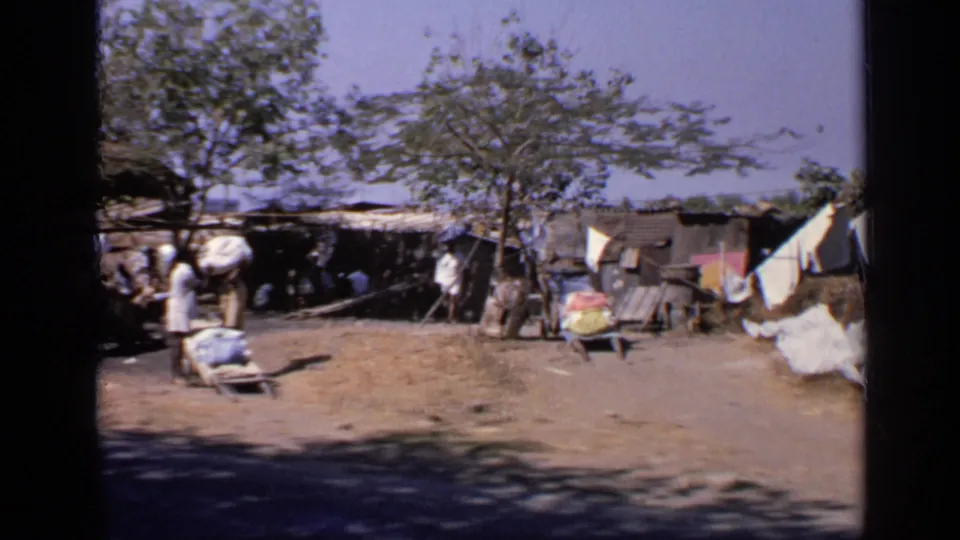 1970 India home-movie frame showing people at a distance near shaded shelters, with laundry spread across a rural dirt compound.