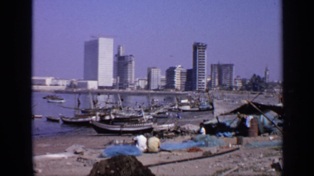 1970 India 8mm frame showing fishing boats near shore, seated fishermen by nets, and high-rise buildings across calm water.