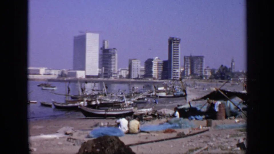 1970 India 8mm frame showing fishing boats near shore, seated fishermen by nets, and high-rise buildings across calm water.