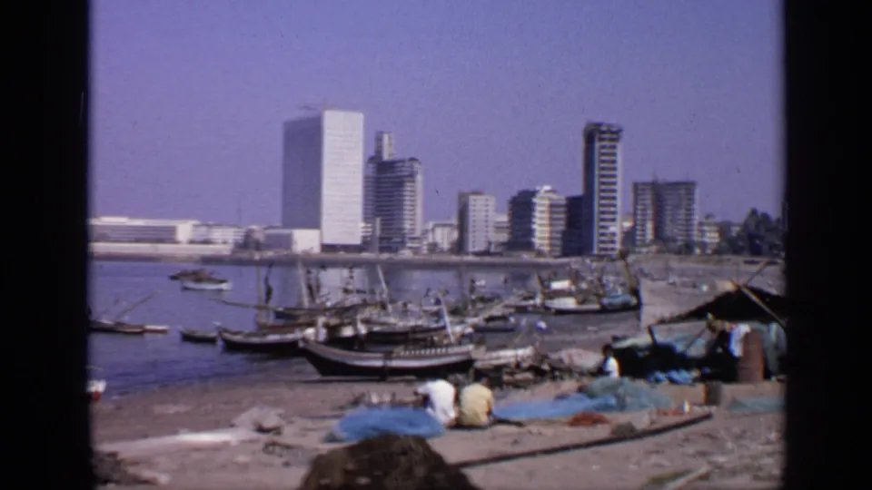 1970 India waterfront view with wooden fishing boats, seated people near blue nets, and a modern city skyline in the background.