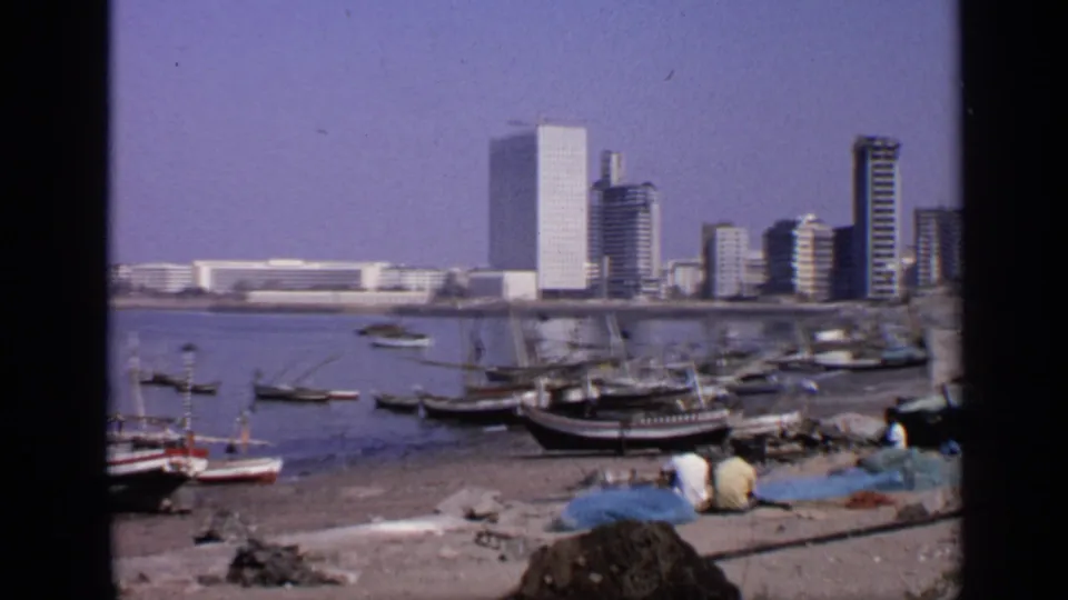 1970 India 8mm frame with fishing boats, blue nets on the shore, seated people, and tall waterfront buildings behind them.