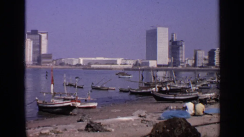 1970 India waterfront frame showing small fishing boats, shoreline workers, calm water, and modern high-rises across the bay.