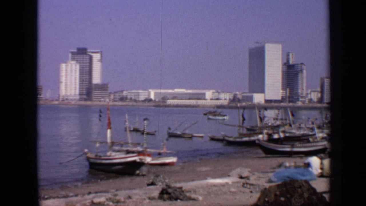 Small boats on a shoreline with calm water and multi-storey buildings across the harbor.
