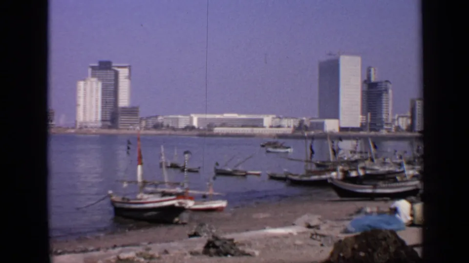 1970 India waterfront view with fishing boats near shore and tall modern buildings across calm harbor water.