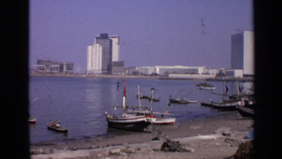 1970 India harbor scene showing moored fishing boats, a sandy shore, and modern buildings across the water.