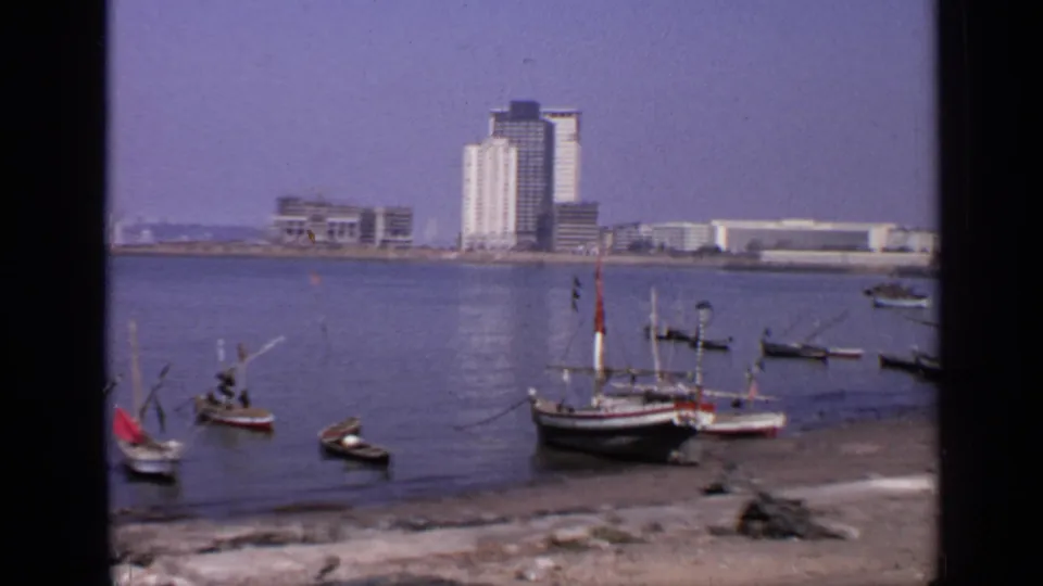 1970 India waterfront footage with several small boats in the foreground and multi-storey buildings in the distance.