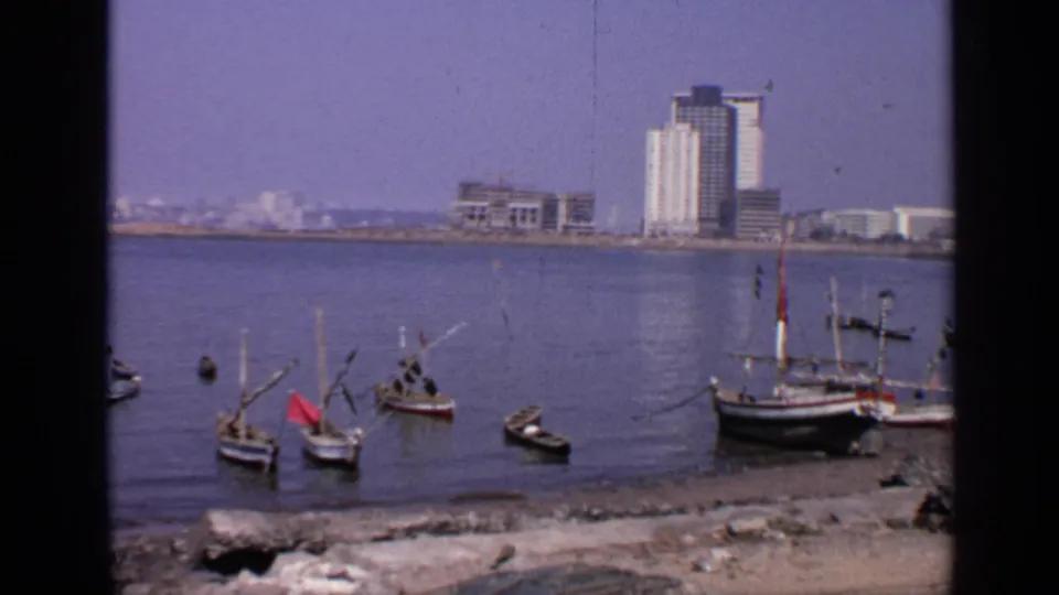 1970 India waterfront view with boats near shore, calm water, and multi-storey buildings beyond the harbor.