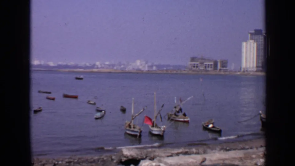 1970 India waterfront scene showing small boats in calm water and a tall modern building on the far shore.