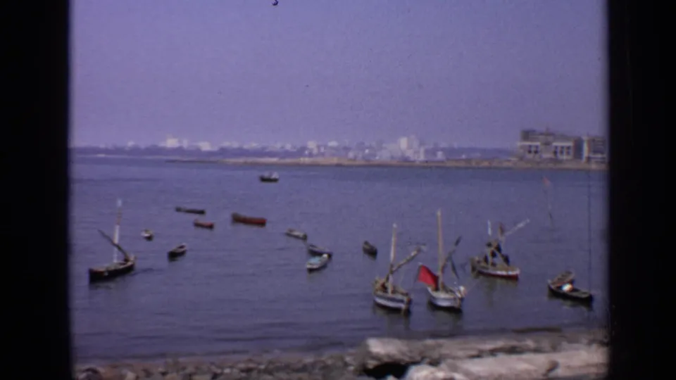 1970 India coastal frame with scattered small boats, low waves, and pale buildings along the far shoreline.