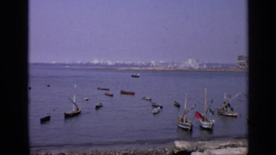 1970 India shoreline view with traditional boats in the foreground water and a distant urban waterfront beyond.