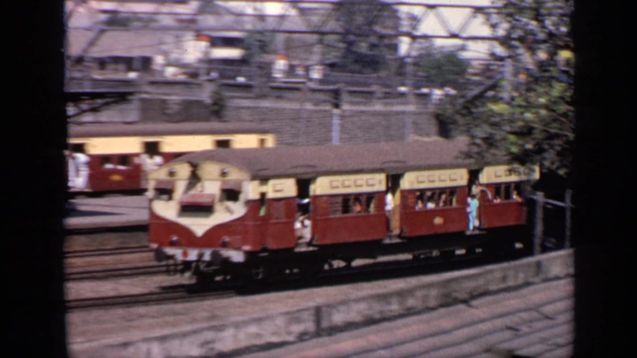 Red-and-cream passenger train passing tracks beside tin-roofed sheds and overhead rail structures.