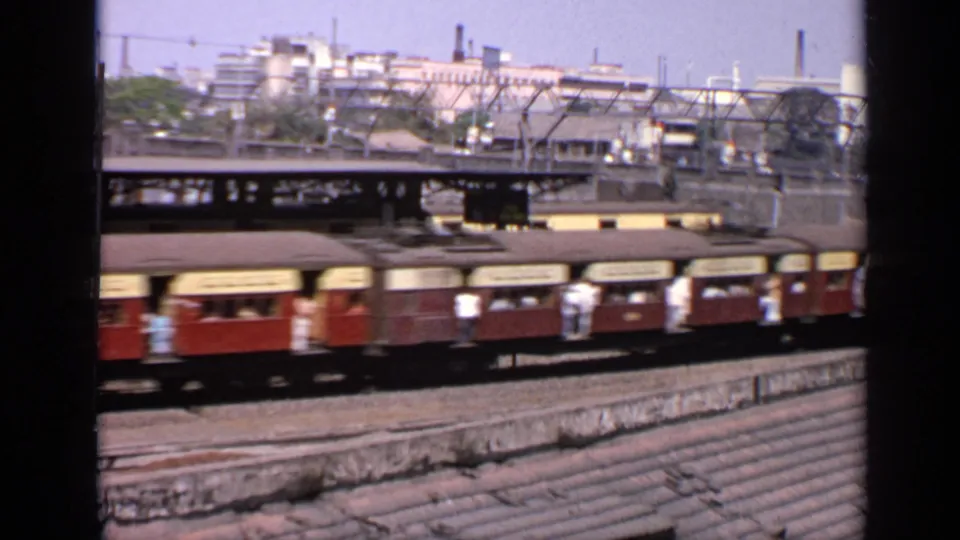 1970 India frame of red-and-cream train carriages passing through an urban rail area, with passengers visible near windows and doors.