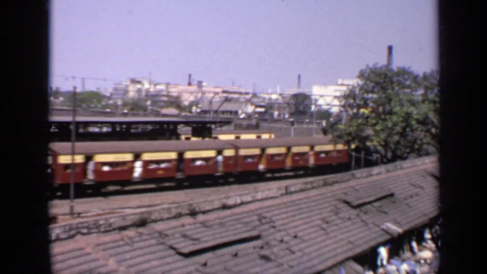 1970 India 8mm frame showing a passing passenger train beyond corrugated trackside roofs, with overhead rail infrastructure in view.