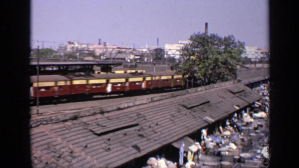 1970 India frame showing the train farther along the tracks, with corrugated roofs, people gathered nearby, and urban rail infrastructure behind.