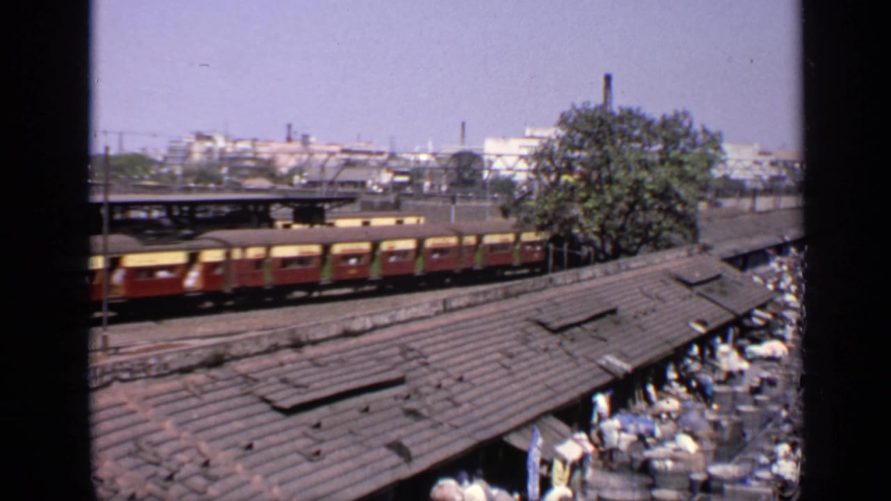 Brown and yellow train passing beside corrugated roofs and a crowded market area.