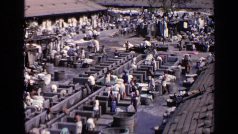 High-angle view of an outdoor India wash center in 1970, with workers moving among concrete basins and cloth piles.
