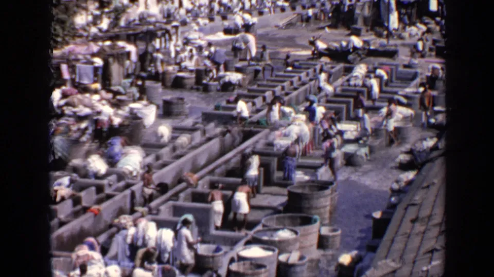 Archival 8mm frame from India showing workers at long concrete washing channels with fabric piles and barrels nearby.
