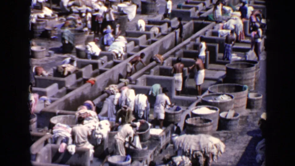 High-angle 1970 India frame showing workers gathered near concrete basins, large barrels, and pale fabric bundles.