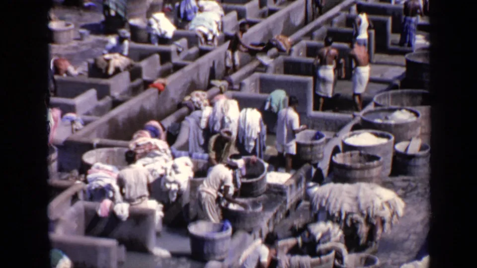 Overhead 1970 India 8mm view of people working among concrete laundry cubicles, tubs, wet basins, and piles of fabric.