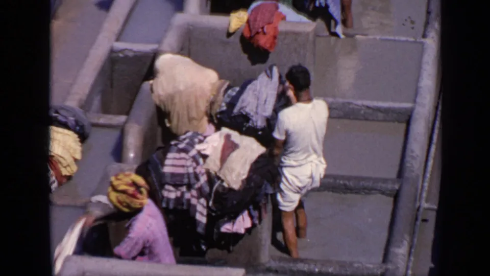 1970 India home-movie frame showing a man in light clothing beside concrete wash cubicles and bundled garments under bright sun.