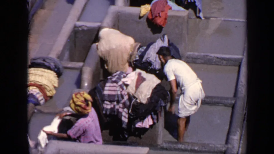 Overhead 1970 India frame of workers near concrete laundry bays, with stacked garments and a person in a colorful head covering visible.