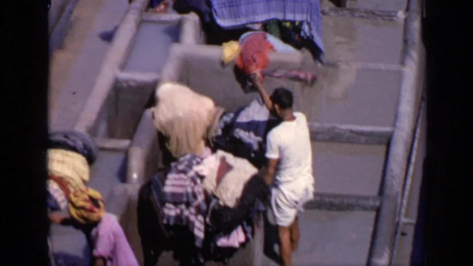 1970 India 8mm still of an outdoor laundry area with a worker, concrete wash bays, draped textiles, and strong sunlight.