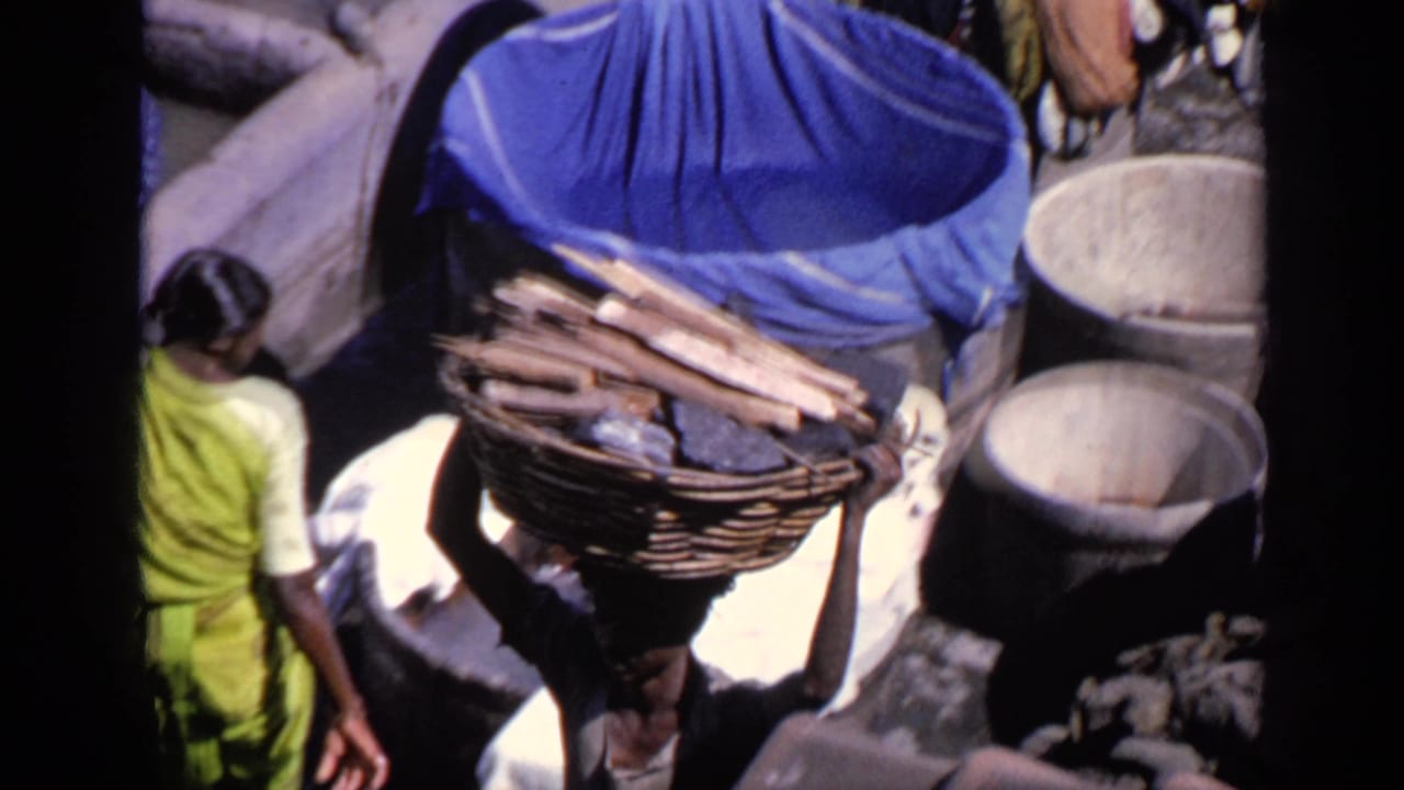 Person carrying a basket of wood on their head in a crowded outdoor area.