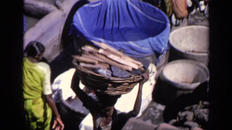1970 India 8mm frame showing a person balancing a basket of wood while others stand near cloth bundles and tubs.