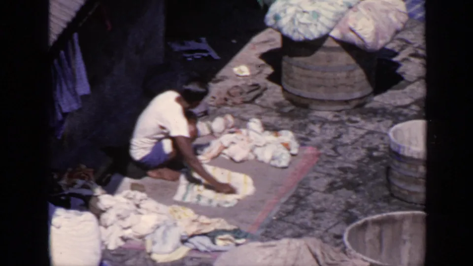 1970 India 8mm frame with a young person crouched beside laundry on a mat, with barrels and fabric bundles nearby.