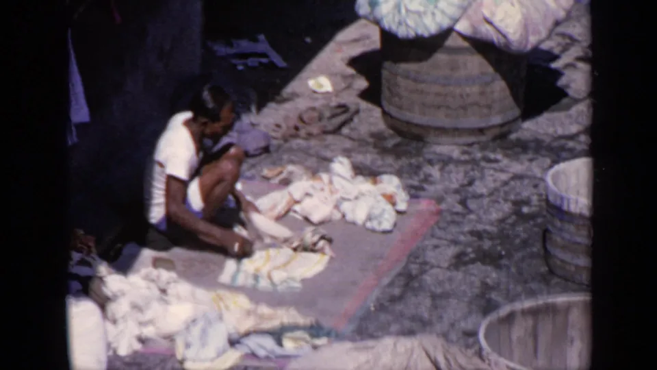 1970 India 8mm frame showing a young person handling laundry on the ground beside cloth piles, tubs, and shaded fabric.