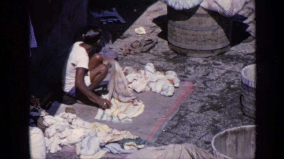 1970 India 8mm frame of a young person lifting fabric beside laundry piles, a mat, and round tubs in daylight.